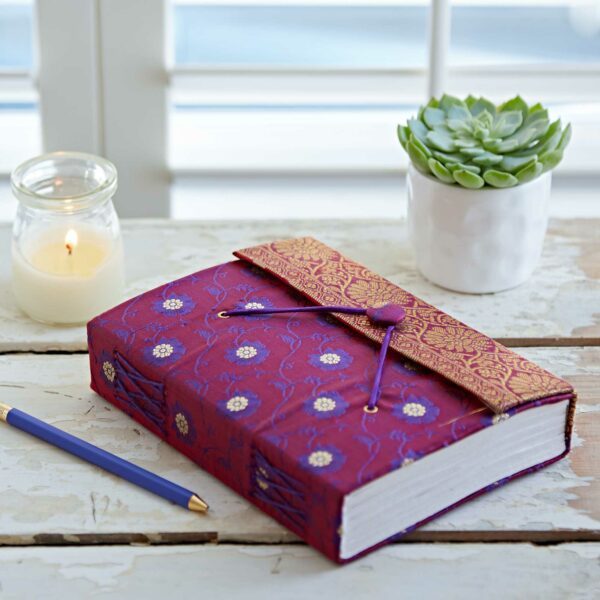 A purple decorative journal with a clasp lies next to a blue pen. A lit candle and a small potted succulent sit nearby on a wooden surface.