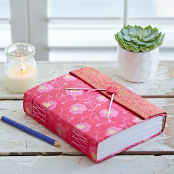 Decorative pink journal with a clasp, a lit candle, a pencil, and a small potted succulent on a white wooden table.