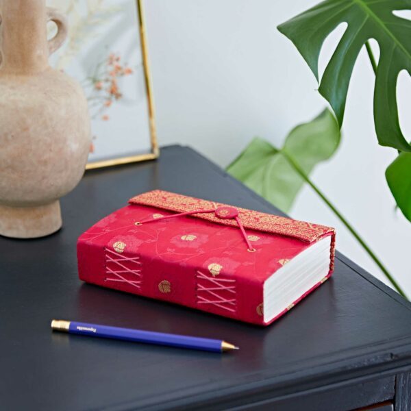 Red journal with decorative cover on a dark table next to a blue pen. Vase and large green leaf in the background.