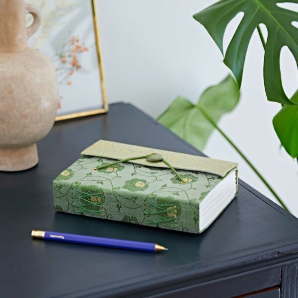 Green embroidered journal on a black wooden desk next to a blue pen. A beige vase and framed artwork are in the background with a large green leaf.