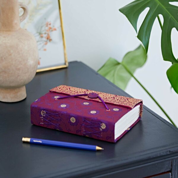 Purple and red floral-patterned journal with tie closure on a black table beside a blue pen and a potted plant.