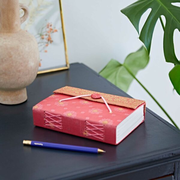 A red fabric-bound notebook with floral patterns and a ribbon clasp lies on a dark table next to a blue pen and a ceramic vase.