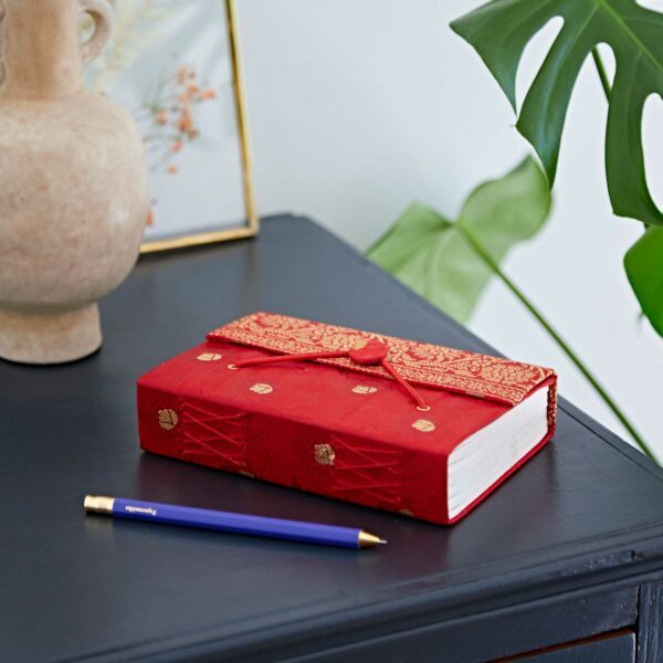 A decorative red journal with gold accents and ribbon closure on a dark surface, next to a blue pen. A ceramic vase and a large green leaf in the background.