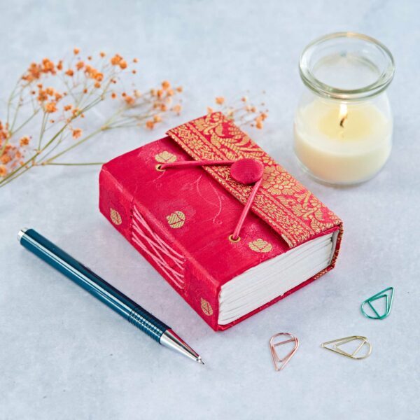 A red fabric-covered journal with a button closure, a lit candle, a blue pen, orange flowers, and three colorful paper clips on a light surface.