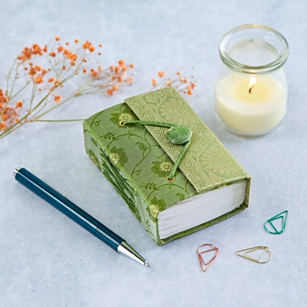 A small green fabric-covered notebook, a blue pen, a lit candle in a jar, dried flowers, and three paper clips on a gray surface.