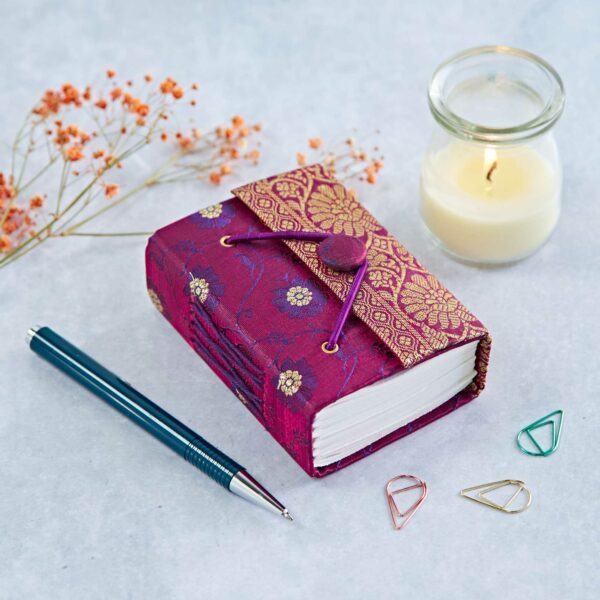 A small, ornate notebook with a decorative cover lies beside a lit candle, a pen, dried flowers, and colorful paper clips on a light surface.