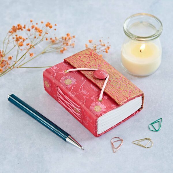 A small red notebook with a floral design, a pen, a lit candle, several paperclips, and orange dried flowers are arranged on a light surface.