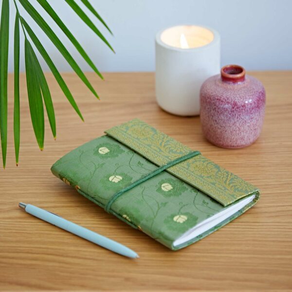 Green journal with a decorative cover, a blue pen, a lit white candle, and a small ceramic vase on a wooden table with a palm leaf nearby.