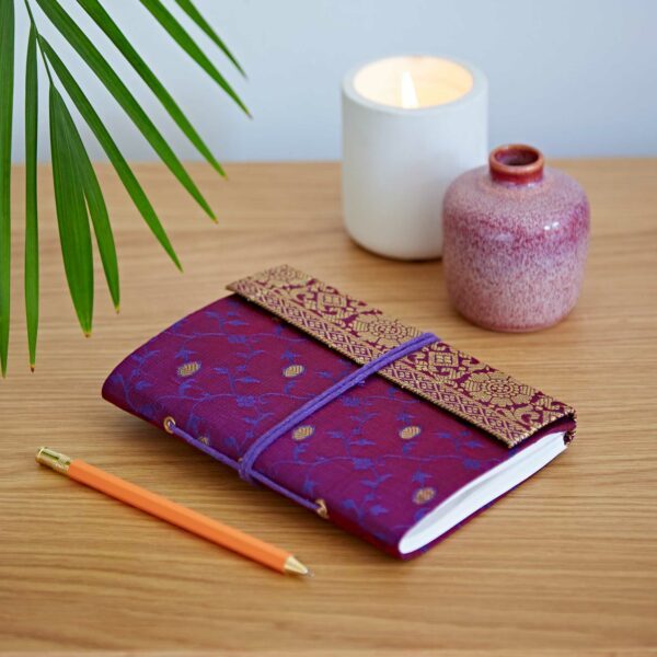 Purple patterned journal and orange pen on wooden table, near a lit candle, pink vase, and green plant.