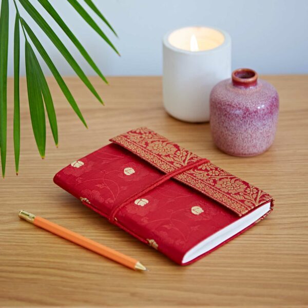 Red fabric-covered notebook with a floral pattern on a wooden table, next to an orange pencil. A lit candle and a small pink vase are in the background. Palm leaves hang above.