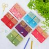 A colorful and inspiring workspace is set with six Mini Sari Fabric Notebooks, each featuring vibrant gold designs, arranged in a grid on a white surface. Accompanying them are two pencils, two paper clips, scissors, and a potted plant.