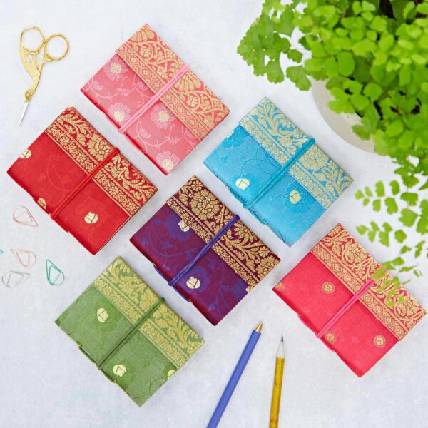 A colorful and inspiring workspace is set with six Mini Sari Fabric Notebooks, each featuring vibrant gold designs, arranged in a grid on a white surface. Accompanying them are two pencils, two paper clips, scissors, and a potted plant.