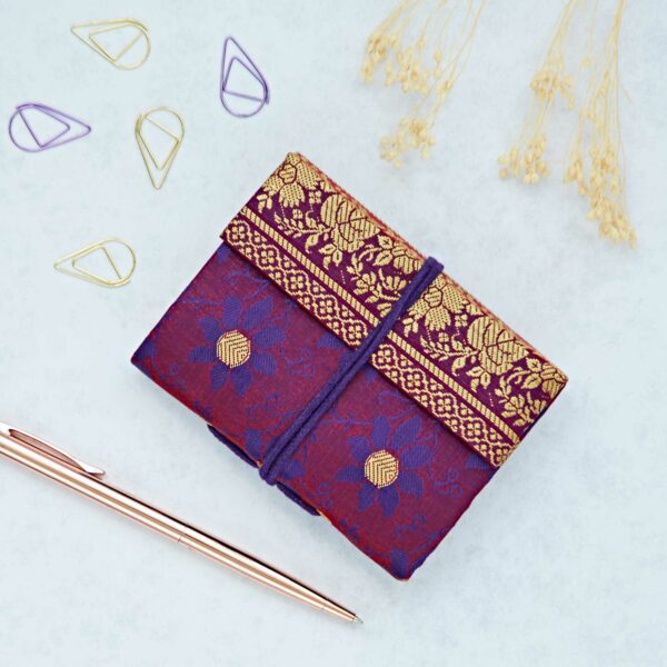 Mini Sari Fabric Notebook featuring a decorative purple and gold design with an intricate floral pattern, complemented by a gold pen, paper clips, and dried flowers on a light background.