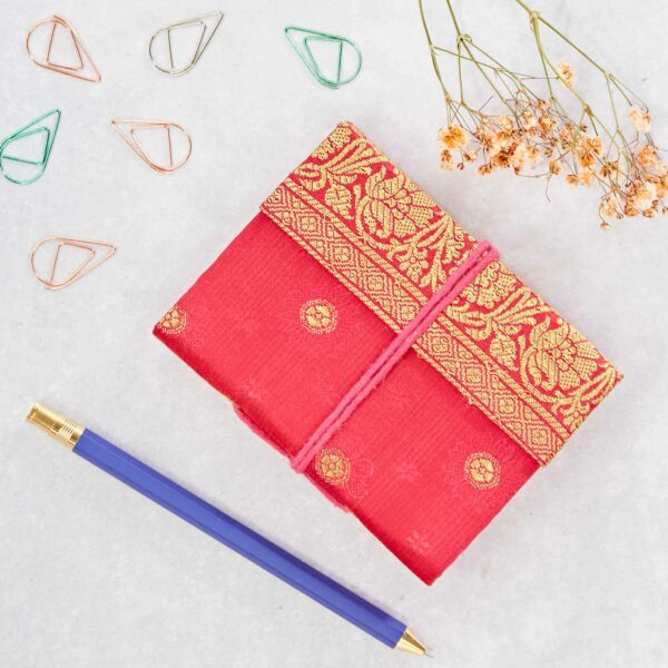 Red fabric-covered notebook with gold patterns, blue pen with gold accents, paper clips, and dried flowers on a light surface.