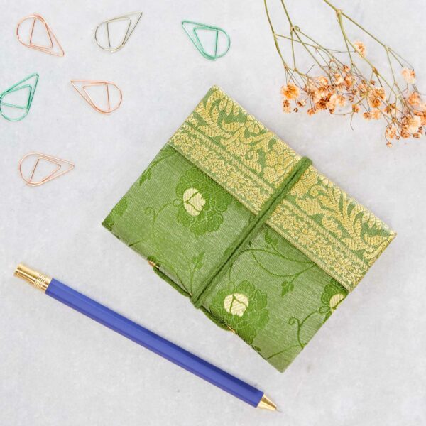 A small green fabric-covered notebook with floral patterns is placed on a surface. Nearby are a blue pen, dried flowers, and paper clips in green and copper colors.