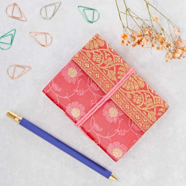 A red and gold patterned wrapped gift is tied with pink string. It's placed on a marble surface with a blue pen, paper clips, and dried flowers nearby.