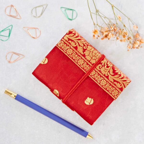 Red and gold patterned journal with a purple pen, dried flowers, and paper clips on a light surface.