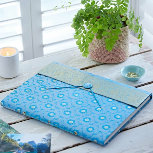 A blue patterned notebook made from sari fabric with a decorative flap lies on a white wooden table beside a potted fern, a lit candle, a small dish, and photographs.