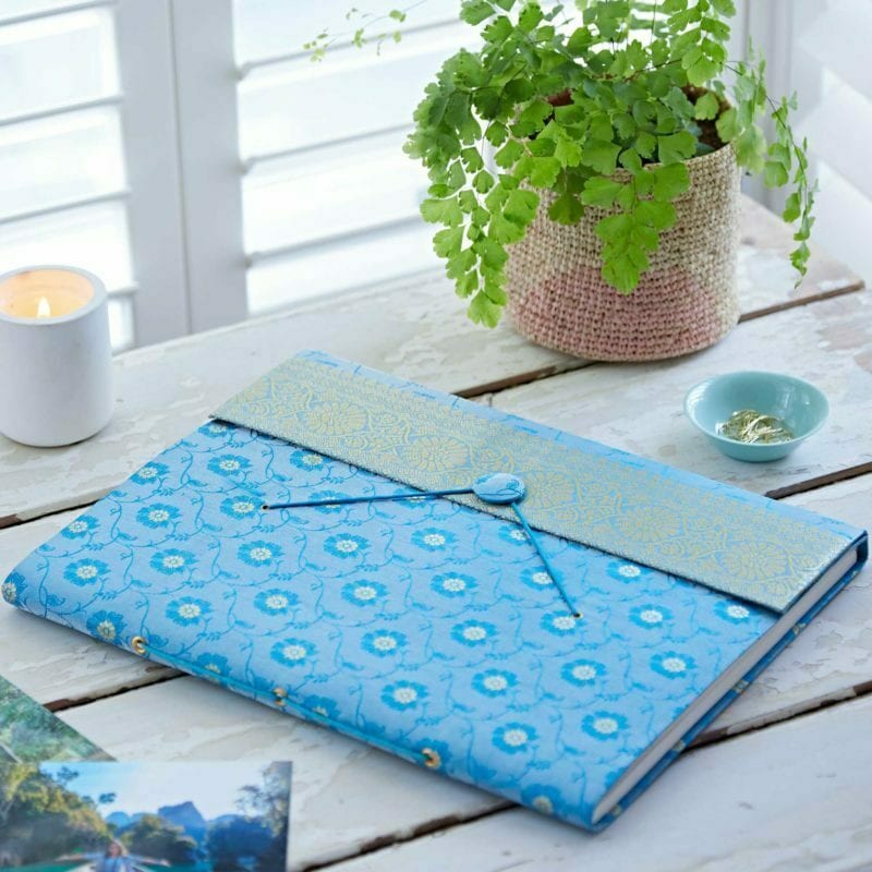 A blue patterned notebook made from sari fabric with a decorative flap lies on a white wooden table beside a potted fern, a lit candle, a small dish, and photographs.