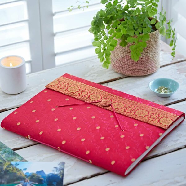 A red and gold patterned notebook made from sari fabric rests on a white wooden table with a green potted plant, a lit candle, a dish with rings, and some photographs—perfect as an extra large photo album or stylish diary.