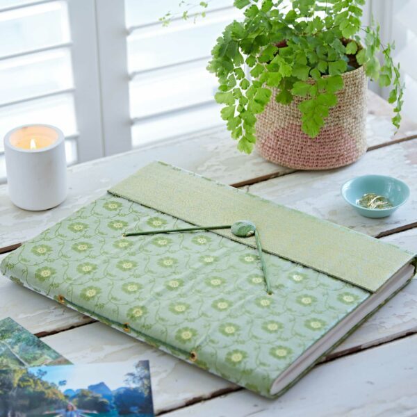 A green floral-patterned Sari Fabric Photo Album rests on a white wooden table beside a potted fern, a lit candle, a small dish with rings, and some photographs.