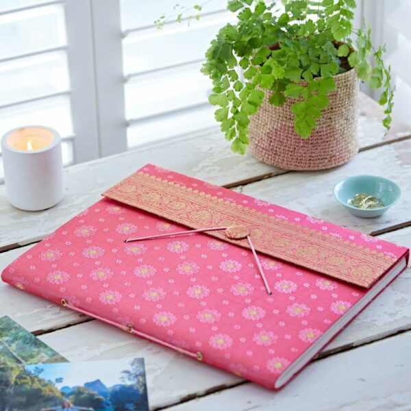 A pink floral-patterned sari fabric photo album with a decorative flap sits on a table near a potted fern, a lit candle, a blue dish with rings, and some landscape photos.