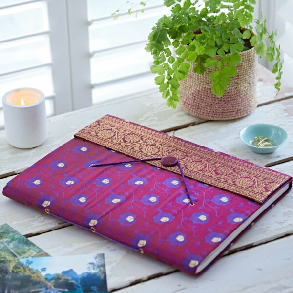 A purple and gold embroidered diary crafted from sari fabric rests on a rustic white table beside a potted fern, a lit candle, a small bowl, and a few holiday photos.