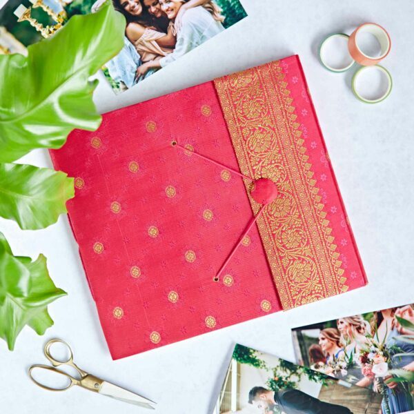 A cerise and gold Sari fabric-covered photo album rests on a white surface, surrounded by photos, scissors, green leaves, and pastel rolls of tape.