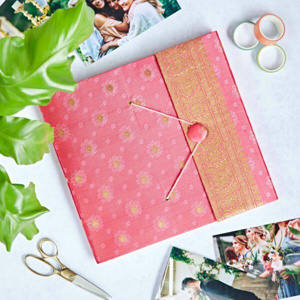 A pink and gold patterned folder inspired by Sari fabric with string fastening lies on a table with photos, scissors, washi tape, and a green plant—perfect for creating your own unique photo album.