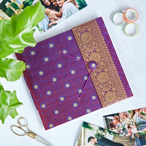 A decorative purple photo album made of sari fabric lies on a table with scattered wedding photos, two rings, a pair of scissors, and a green plant nearby.