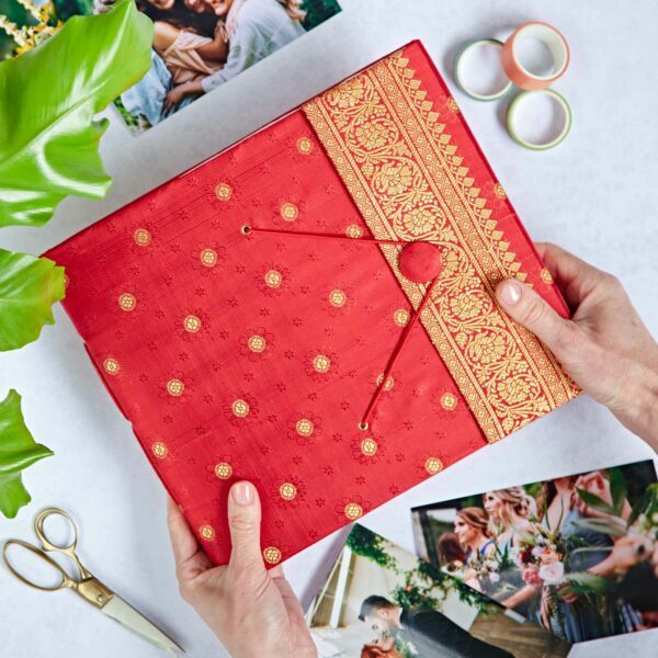 Person holding a gift wrapped in luxurious red sari fabric, surrounded by wedding photos, a photo album, green plant, scissors, and decorative tape on a white surface.