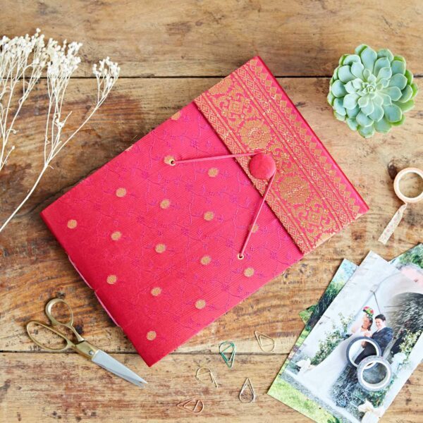 A red and gold patterned notebook, inspired by sari fabric, rests on a wooden table beside dried flowers, a succulent plant, scissors, paperclips, tape, and printed photographs.