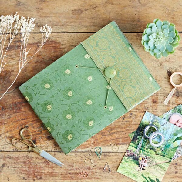 Green floral-patterned folder, resembling a Sari fabric photo album with button fastening, sits on a wooden table surrounded by scissors, dried flowers, succulent, paper clips, washi tape, and printed photos.