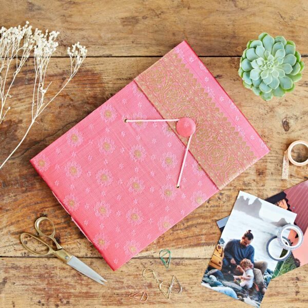 A pink sari fabric embroidered envelope lies on a wooden table with scissors, paper clips, dried flowers, a succulent, washi tape, and scattered photographs—like the start of a unique photo album.