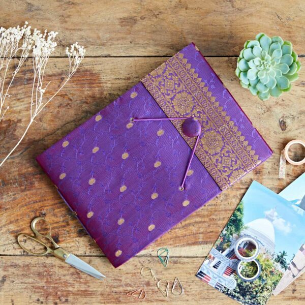 A purple and gold Sari fabric photo album folder rests on a wooden table, surrounded by scissors, paper clips, dried flowers, a succulent plant, washi tape, and postcards.