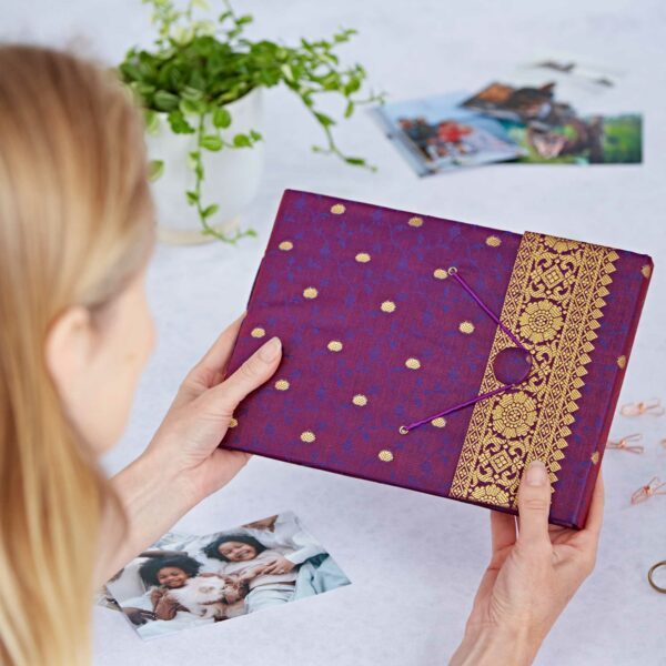 Someone holds a Medium Sari Fabric Photo Album in purple and gold; printed photos and a potted plant rest on the table.