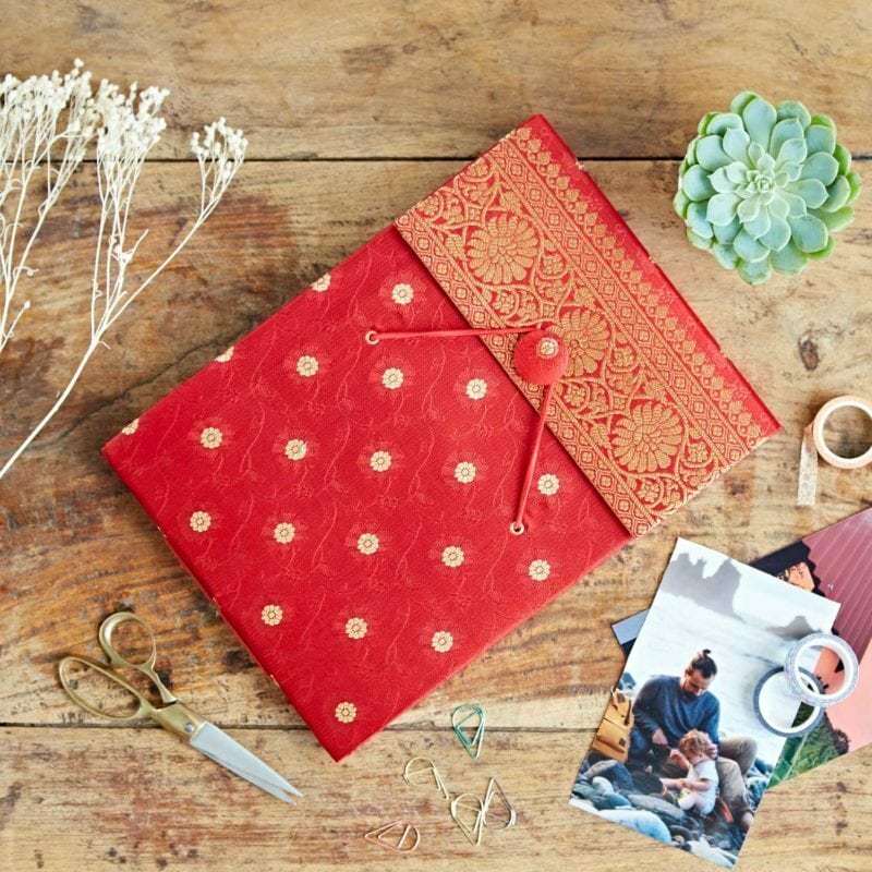 A red and gold decorative photo album on a wooden table, surrounded by scissors, paper clips, a plant, dried flowers, tape, and printed photos—its cover inspired by traditional Sari fabric for an elegant touch.