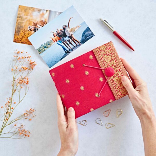 A person holds a cerise patterned diary reminiscent of sari fabric; nearby are two printed photos, a pen, paper clips, and dried flowers on a white surface.