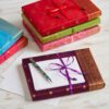 Vibrant sari-inspired notebooks with ribbon closures are stacked on a light wooden surface. On top, an envelope and striped pen from the Sari Letter Writing Set await your letter writing.
