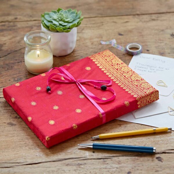 A red and gold wrapped gift with a pink bow on a wooden table, surrounded by a lit candle, a succulent plant, two pens, and handwritten letters.