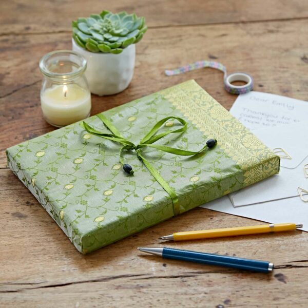 A gift wrapped in green patterned paper with a ribbon on a wooden table, accompanied by a lit candle, a potted plant, a handwritten letter, and two pens.