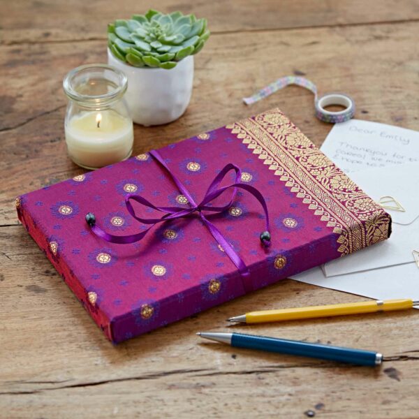 A purple and gold fabric-covered journal with a tie sits on a wooden table, surrounded by a lit candle, a succulent plant, pens, a letter, and decorative tape.
