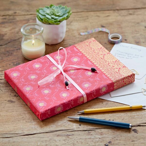 Gift wrapped in red and gold floral paper with ribbon on a wooden table, accompanied by a candle, succulent, letter, tape, pen, and pencil.