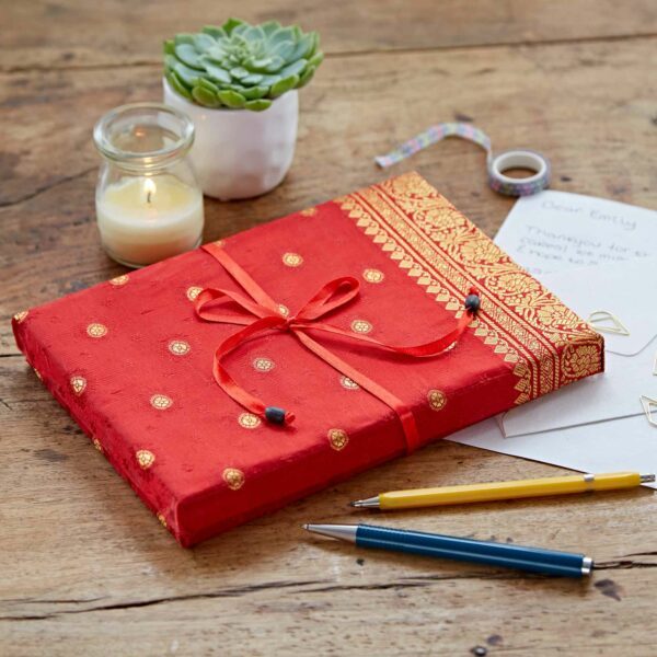 A red, patterned gift box with a ribbon on a wooden table, surrounded by a candle, succulent, pens, and a letter.