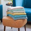 A stack of five Chevron Recycled Cotton Throws in assorted pastel hues rests folded on an orange stool beside a blue armchair.
