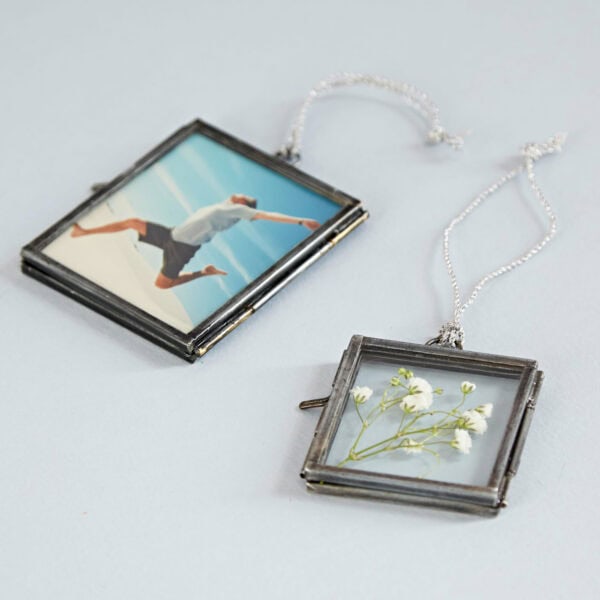 Two Alia Square Glass Hanging Photo Frames lie on a gray surface. The left frame displays a photo of a man jumping on a beach, while the right frame showcases a pressed white flower, reminiscent of capturing treasured moments.