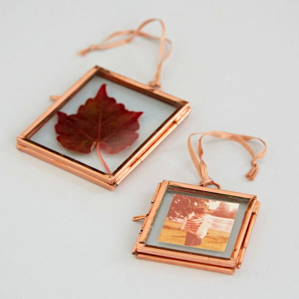 Two Alia Square Glass Hanging Photo Frames with copper frames and ribbons. One frame contains a red autumn leaf, while the other displays a photograph of a person standing outdoors. Both items are placed on a light-colored surface.