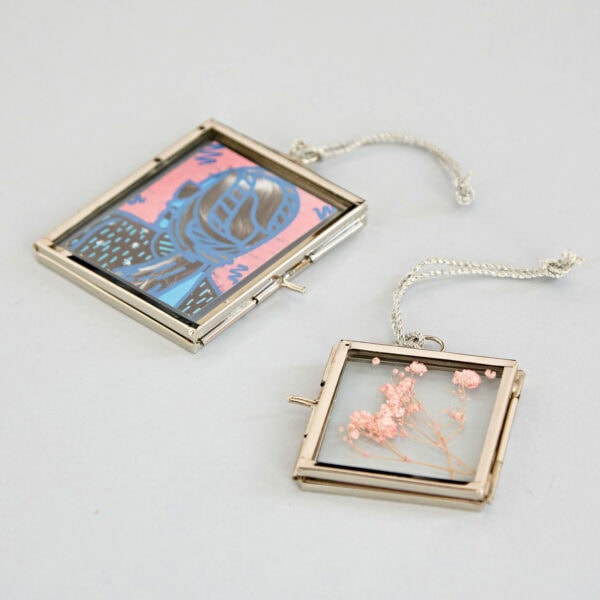 Two Alia Square Glass Hanging Photo Frames on a light grey background resemble elegant square lockets; one contains an illustrated image of a blue knight on a pink background, and the other holds small dried pink flowers.