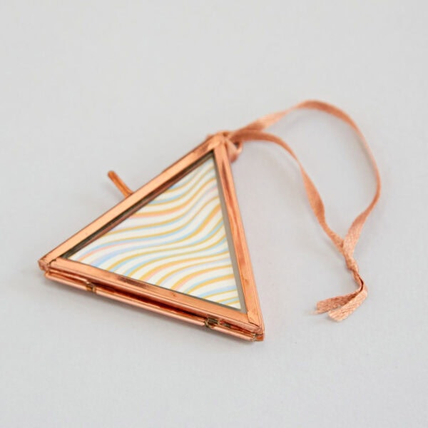 An Alia Mini Triangular Glass Hanging Photo Frame in rose-gold with a wavy, multicolored design inside and a matching ribbon attached.