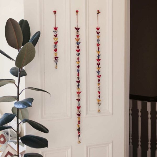 Three strings of colorful decorative hanging ornaments, including the charming Prosperity Bird Bell Tota, are displayed on a white wall next to a large leafy plant, symbolizing prosperity.